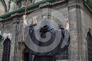 A monk with a staff in a black robe stands in front of an old gloomy building with his staff raised high, light and fire radiates