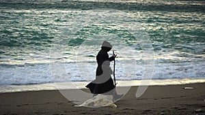Monks wearing black robes walks on the beach with woden stick in hands at morning. Unrecognizable persone, slow motion.
