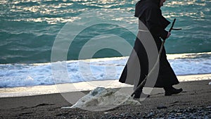 Monks wearing black robes walks on the beach with woden stick in hands at morning. Unrecognizable persone, slow motion.