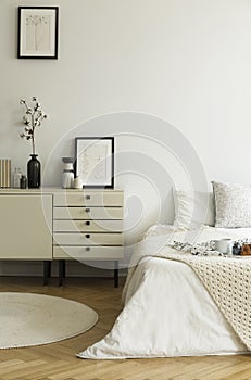 A monochromatic beige and white bedroom interior with a view at a bed and a drawer cabinet standing on a wooden floor. Real photo.