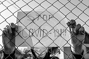 Monochrome image of a desperate teenage boy wearing surgical mask holding a message  behind a wired fence in isolation