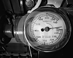 Monochrome image of a large old metal tachometer with a round gauge with the dial marked in numbers mounted on a large engine
