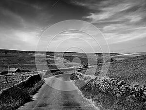 Monochrome image of a narrow country lane surrounded by dy stone walls and dramatic clouds in a sunlit rural pennine landscape on