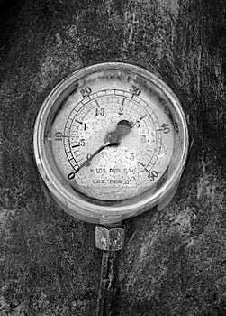 Monochrome image of an round industrial pressure gauge with numbers round the dial mounted on a metal surface