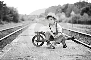 Monochrome portrait of cute boy wearing in retro style sitting on railway station and is waiting for something