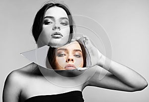 Monochrome portrait of female with mirror shard in hand posing on gray background. Color face reflection in mirror splinter