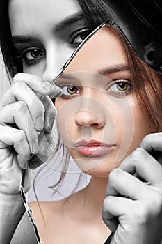 Monochrome portrait of female with mirror shard in hand posing on gray background. Color face reflection in mirror splinter