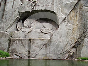 Monument `The Dying Lion` in Lucerne. Switzerland. High relief carved in a steep rock behind an oval pond. The dying lion lies wit