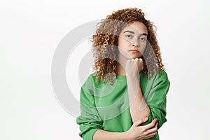 Moody and bored curly girl sulking, lean head on fist and staring with bothered, annoyed face expression, standing