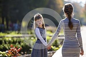 Moody cute child girl holding mother hand looking back on warm day outdoors. Family relationship and recreation