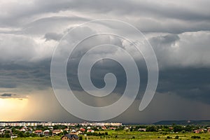 Moody landscape with dark stormy clouds with falling heavy downpour shower rain over distant town buildings in summer