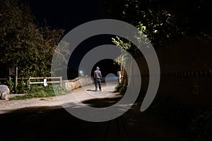 A moody night scene of a hooded man standing on a country lane lit up with a street light on a summers night