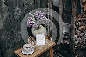 Moody rustic spring outdoor still life. Purple, white lilac flowers bouquet in textured vase. Cup of coffee, books