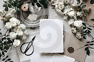 Moody wedding table mockup scene. Stationery composition with fading white rose flowers, silver plate, black scissors