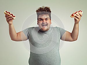 More is better. Studio shot of a happy overweight man holding 2 hotdogs in the air.