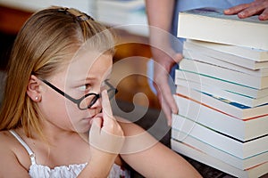 More. A cute little blonde girl looking worried as someone brings her a stack of books in a library.