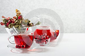 Morning autumn teatime with red herbal tea in transparent tea pot, cup with hawthorn, rosehip berry, dried leaves in sun beam.