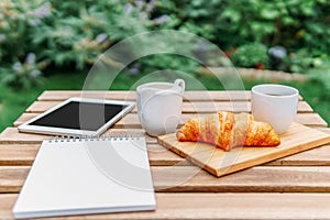 Morning Breakfast In Green Garden With French Croissant, Coffee Cup, Orange Juice, Tablet and Notes Book On Wood Table