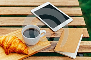 Morning Breakfast In Green Garden With French Croissant, Coffee Cup, Orange Juice, Tablet and Notes Book On Wood Table