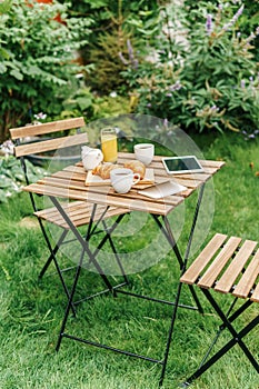 Morning Breakfast In Green Garden With French Croissant, Coffee Cup, Orange Juice, Tablet and Notes Book On Wood Table