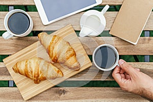 Morning Breakfast In Green Garden With French Croissant, Coffee Cup, Orange Juice, Tablet and Notes Book On Wood Table