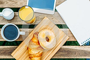 Morning Breakfast In Green Garden With French Croissant, Coffee Cup, Orange Juice, Tablet and Notes Book On Wood Table