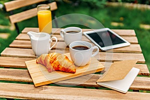 Morning Breakfast In Green Garden With French Croissant, Coffee Cup, Orange Juice, Tablet and Notes Book On Wood Table