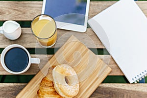 Morning Breakfast In Green Garden With French Croissant, Coffee Cup, Orange Juice, Tablet and Notes Book On Wood Table