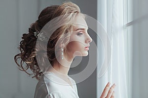 Morning of the bride. Beautiful young woman in elegant white robe with fashion wedding hairstyle standing near the