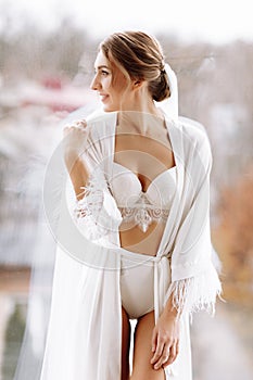 Morning of bride before the wedding. beautiful young woman with long veil in a white robe and a negligee at the window