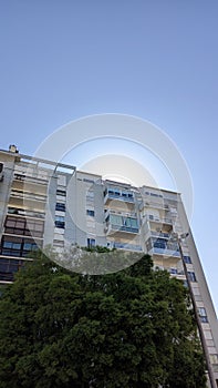 In the morning, a bright sun appears from behind a high-rise apartment building against a blue sky