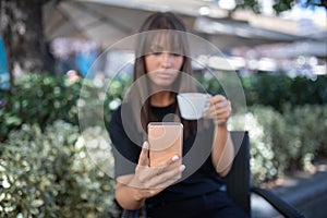 Morning in cafe - attractive woman in black drinking coffee and make selfie photo for social networks. Focus on phone