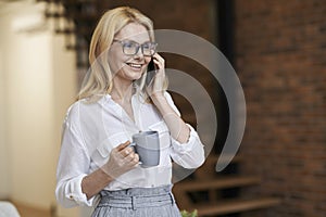 Morning. Cheerful middle aged woman with blonde hair and glasses holding a cup of coffee or tea while talking on her