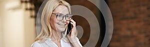 Morning. Cheerful middle aged woman with blonde hair and glasses holding a cup of coffee or tea while talking on her