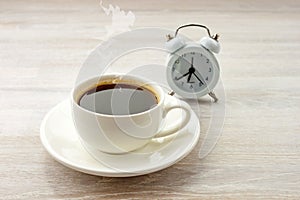 Morning coffee black hot espresso with steam in white cup, saucer on wooden table with clock
