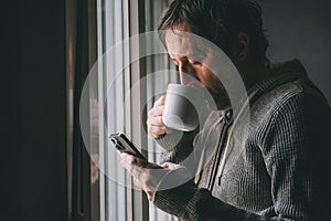 Morning coffee break, adult caucasian unkempt man drinking coffee from white mug and browsing through social media feed on his