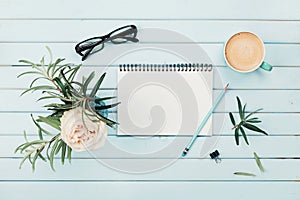 Morning coffee cup, clean notebook, pencil, eyeglasses and vintage rose flower in vase on blue rustic desk overhead view.