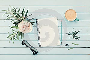 Morning coffee cup, clean notebook, pencil, eyeglasses and vintage rose flower in vase on blue rustic table top view. Flat lay.