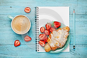 Morning coffee cup with fresh croissant, strawberry and empty notebook on turquoise rustic table from above, tasty breakfast