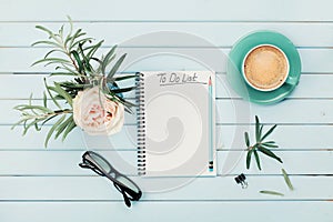 Morning coffee cup, notebook with to do list, pencil, eyeglasses and vintage rose flower in vase on blue rustic table from above.