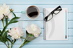 Morning coffee mug, empty notebook, pencil, glasses and white peony flowers on blue wooden table, cozy summer breakfast