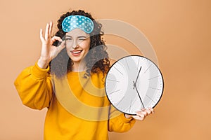 Morning concept. Beautiful curly young woman posing over beige background with clock and sleeping mask. Ok sign