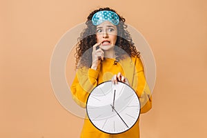 Morning concept. Beautiful shocked amazed curly young woman posing over beige background with clock and sleeping mask
