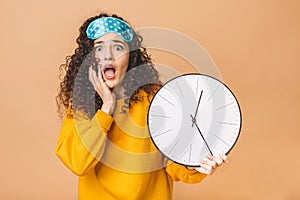 Morning concept. Beautiful shocked amazed curly young woman posing over beige background with clock and sleeping mask