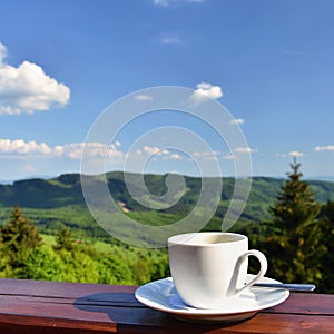 Morning cup of coffee with a beautiful mountain landscape background. White cup and saucer - espresso on a wooden table.