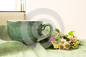 Morning herbal tea cup and summer flowers near window view on blue green cotton napkin background