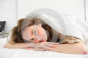 Morning in hotel room. Young smiling woman lying in bed, wake up, eyes opened, looking relaxed and happy