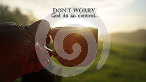 Morning inspirational quote - Don't worry. God is in control. With person holding coffe or tea cup against the warm sunrise