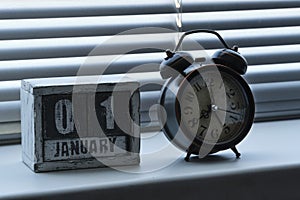 Morning of January 01 on wooden calendar standing window with blinds next to an alarm clock showing eight o clock.