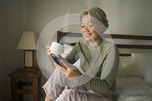 Morning lifestyle portrait of attractive and happy middle aged woman on her 50s having coffee on bed using internet mobile phone